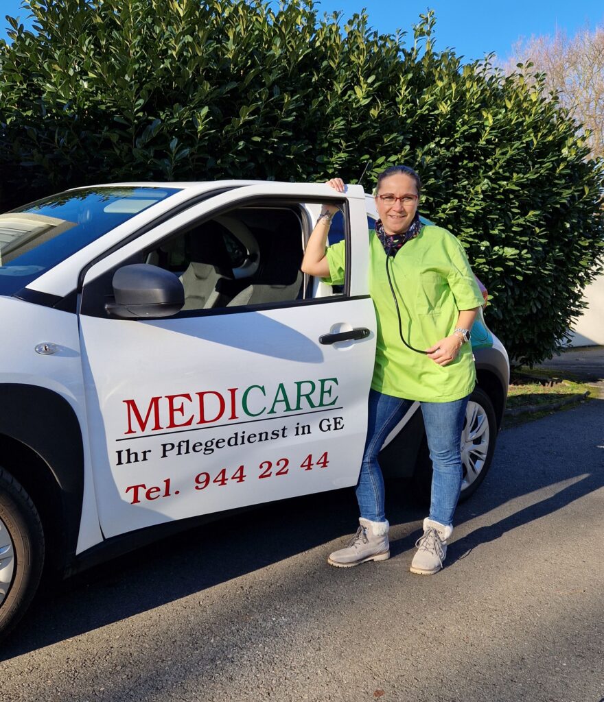 Person in grünem Shirt lächelt neben weißem Auto mit MEDICARE-Logo, blauer Jeans, hellem Schuhwerk.